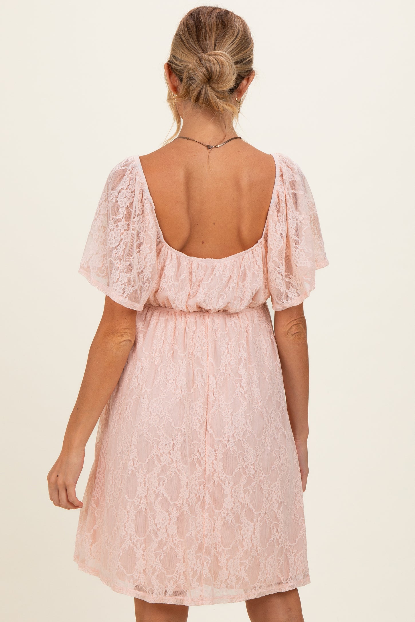 woman wearing a light pink lace maternity dress with flutter sleeves and a loose fit, showing the back view