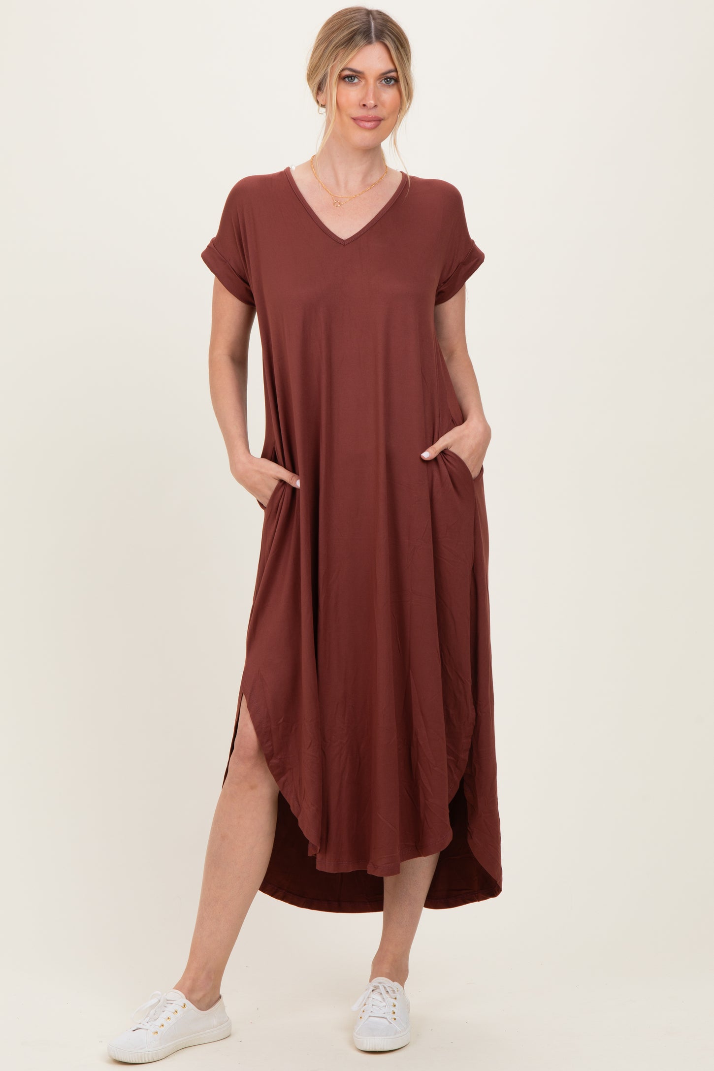woman wearing a rust-colored v-neck short sleeve maxi dress with pockets and high-low hem, standing in white sneakers