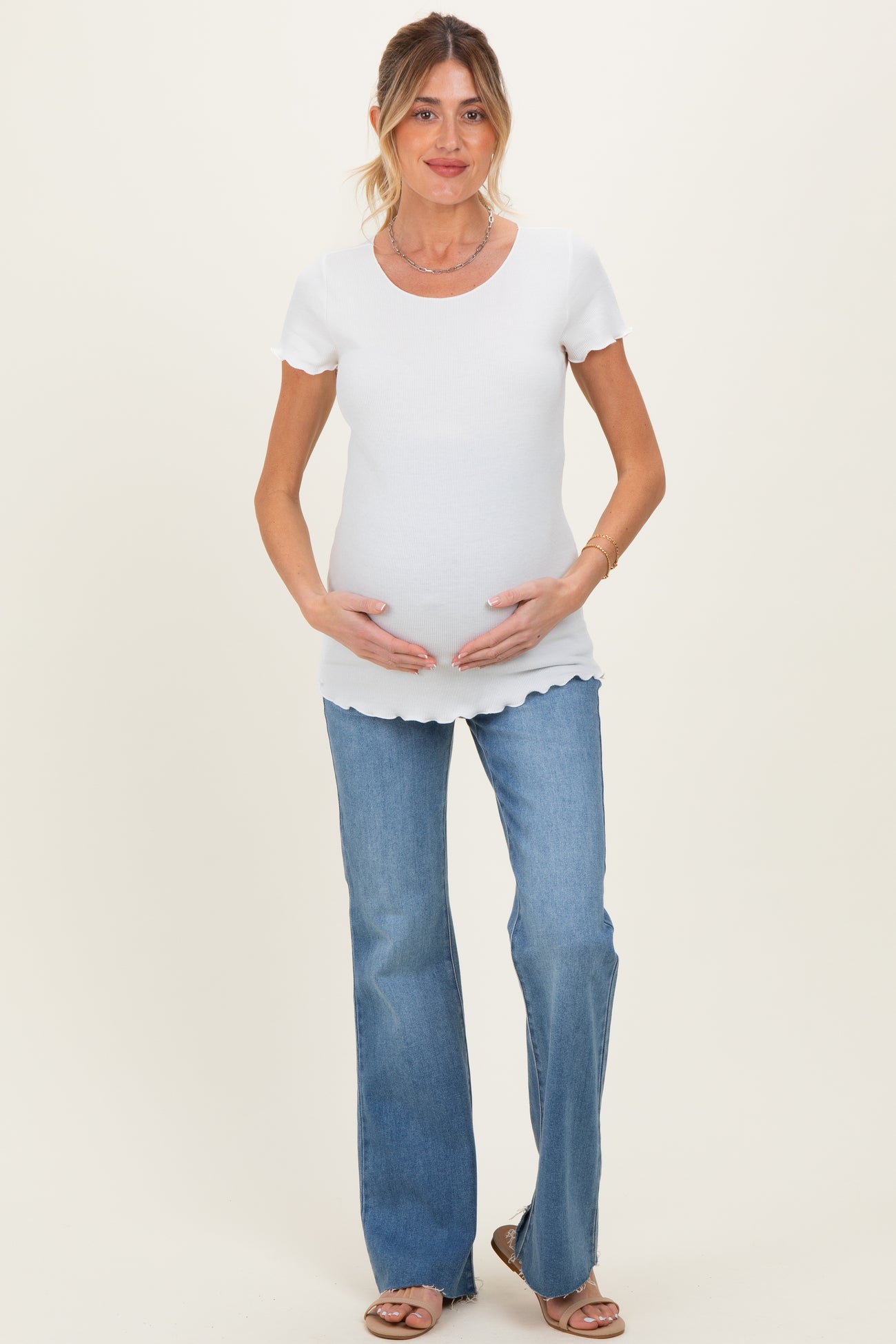 woman wearing a white ribbed maternity top and holding her baby bump, paired with light blue flared jeans