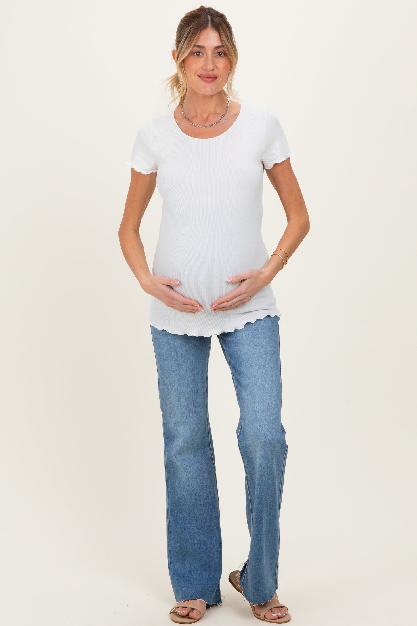 woman wearing a white ribbed maternity top and holding her baby bump, paired with light blue flared jeans