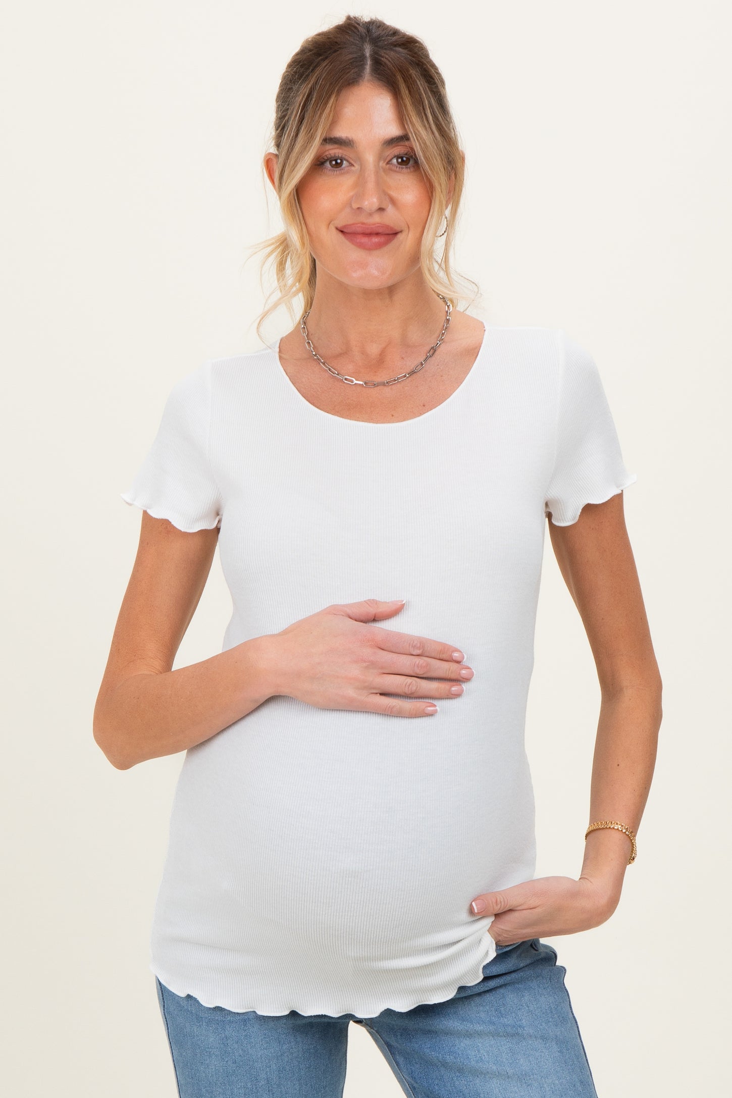 woman wearing a white short sleeve maternity top with scalloped details and holding her baby bump
