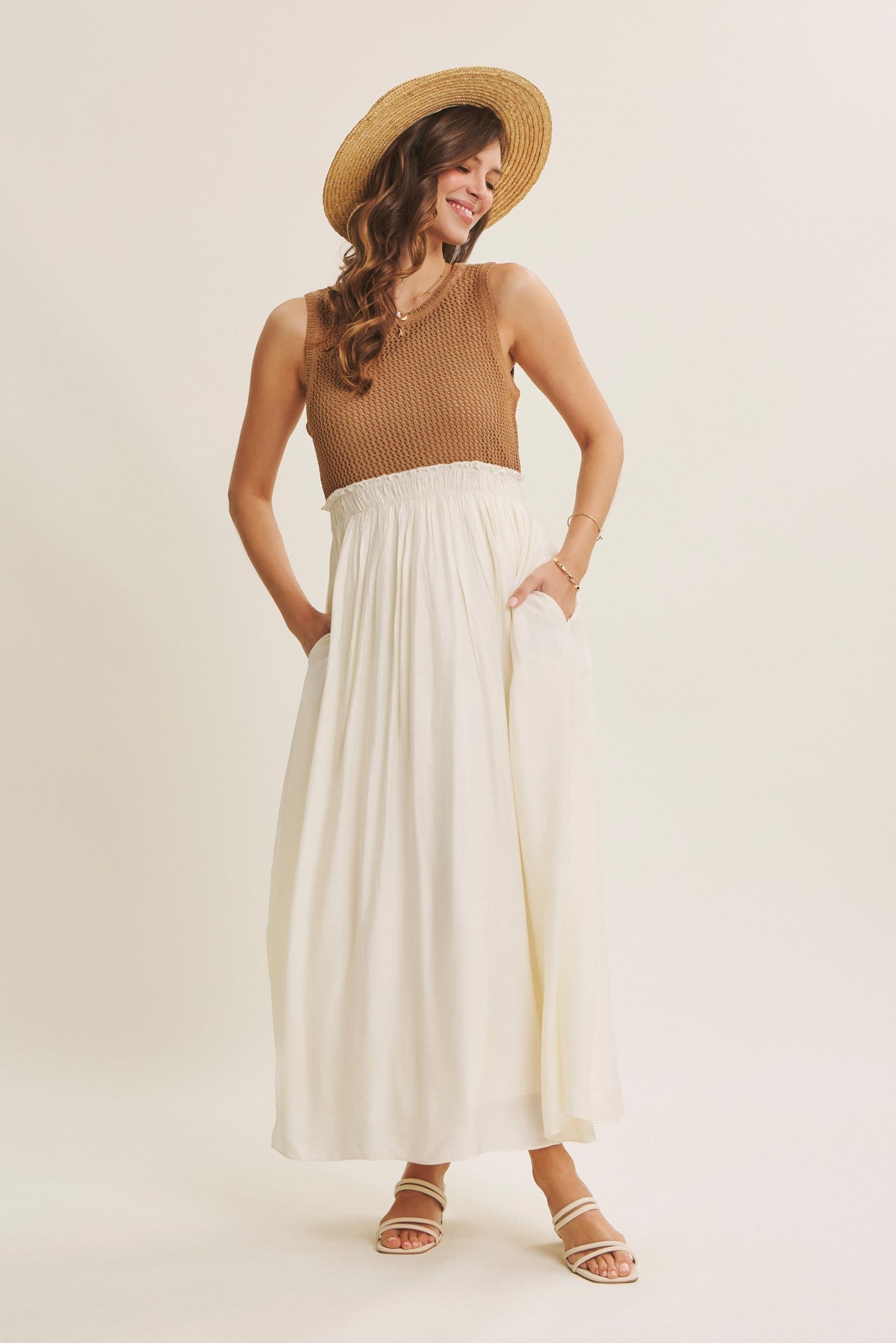 woman wearing a tan knit tank top and a flowy white maxi skirt, smiling and posing with her hands in pockets, wearing a straw hat