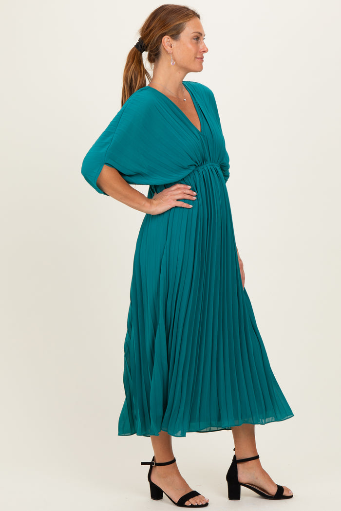 woman wearing a teal pleated midi dress with a deep v-neck and batwing sleeves, styled with black heeled sandals