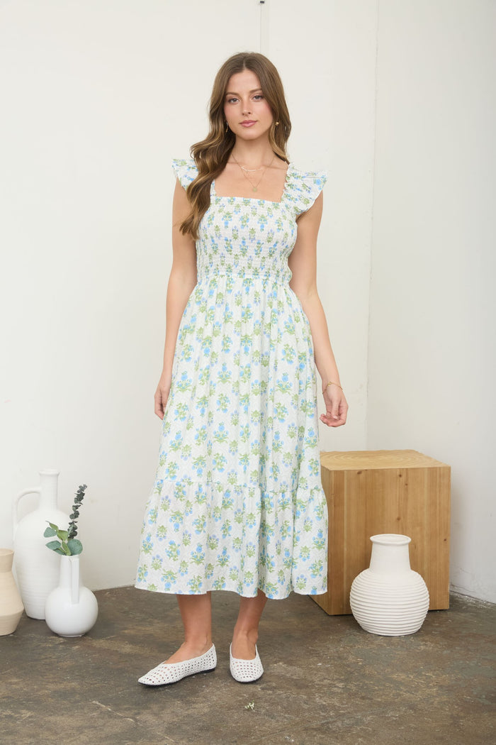 woman wearing a light blue floral maxi dress with ruffled sleeves and a fitted bodice, standing on a wooden floor