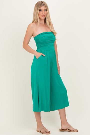 woman wearing a strapless teal jumpsuit with a cinched waist and pockets, standing and smiling