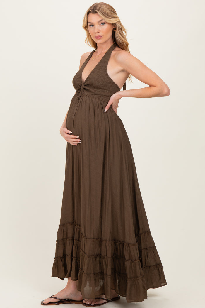 woman wearing a brown maternity maxi dress with a halter neckline and a tiered ruffle hem, holding her baby bump