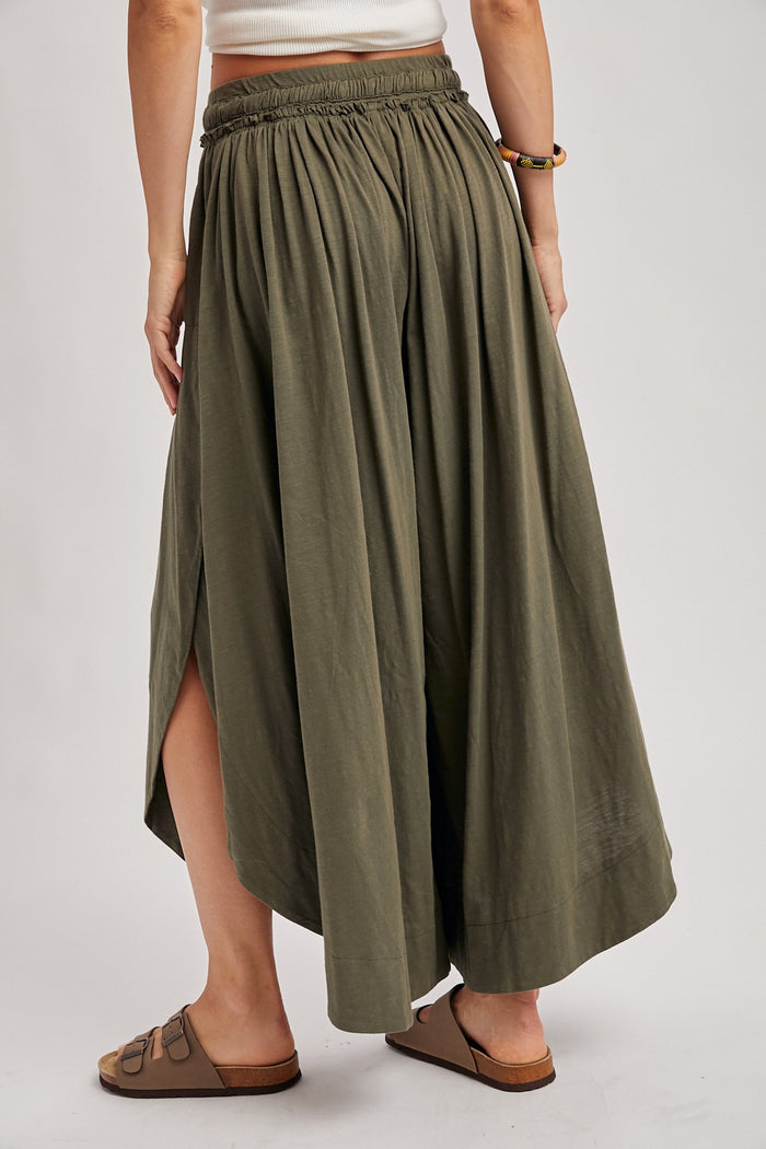 woman wearing a long olive green maxi skirt with a pleated design and side slits, showing the back view