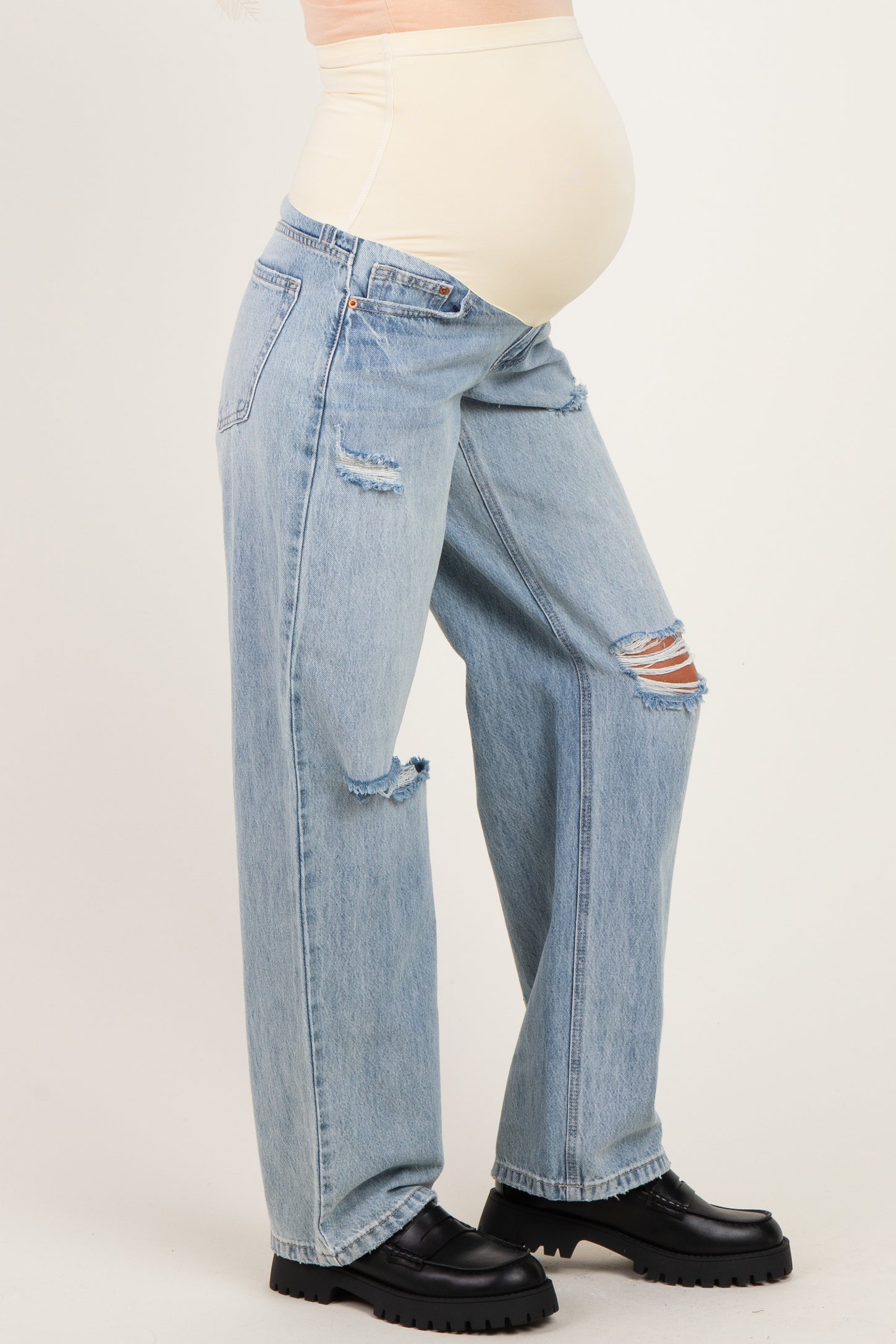 woman wearing light blue distressed maternity jeans and a beige belly support top, showcasing her baby bump
