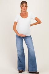 woman wearing a white short sleeve maternity top and blue flared jeans, cradling her baby bump