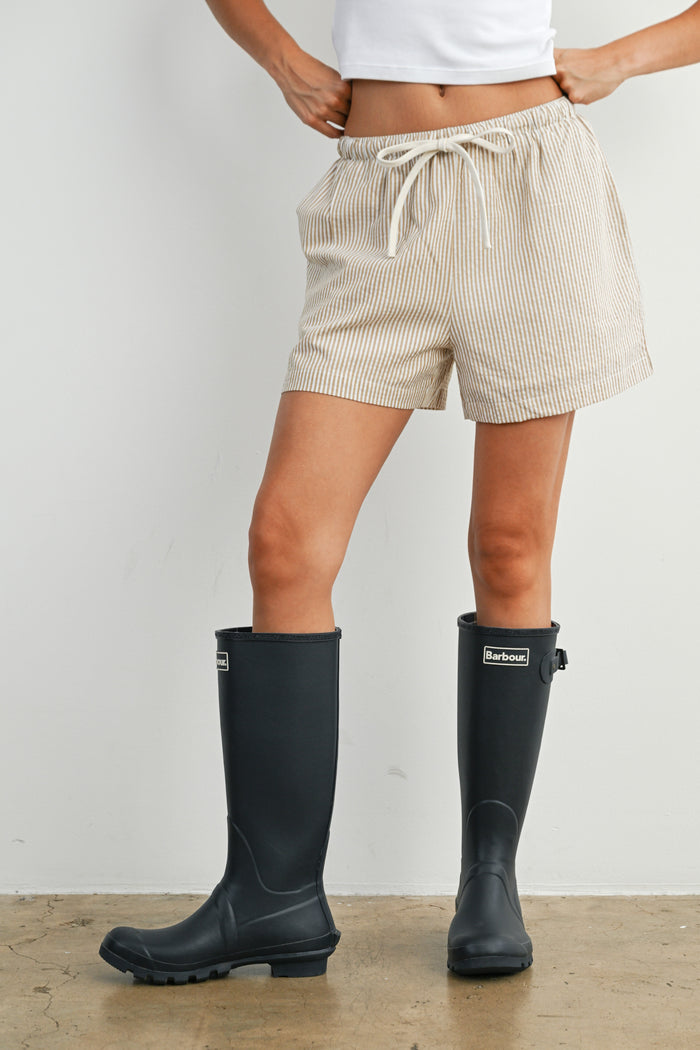 woman wearing beige striped shorts and black rubber rain boots, standing on a textured floor