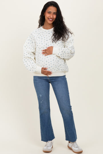 woman wearing a white floral print maternity sweatshirt and blue jeans, holding her baby bump