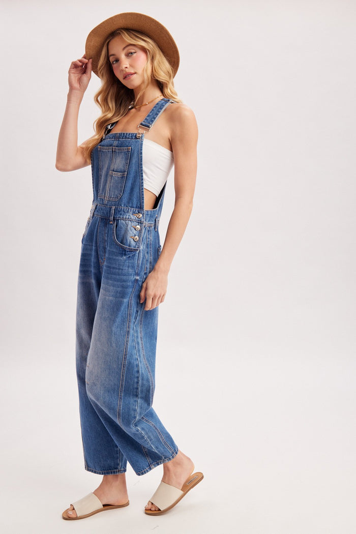 woman wearing a light wash denim overall with wide legs, paired with a white tank top and straw hat