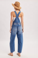 woman wearing blue denim overalls with a relaxed fit, back view, accessorized with a tan straw hat