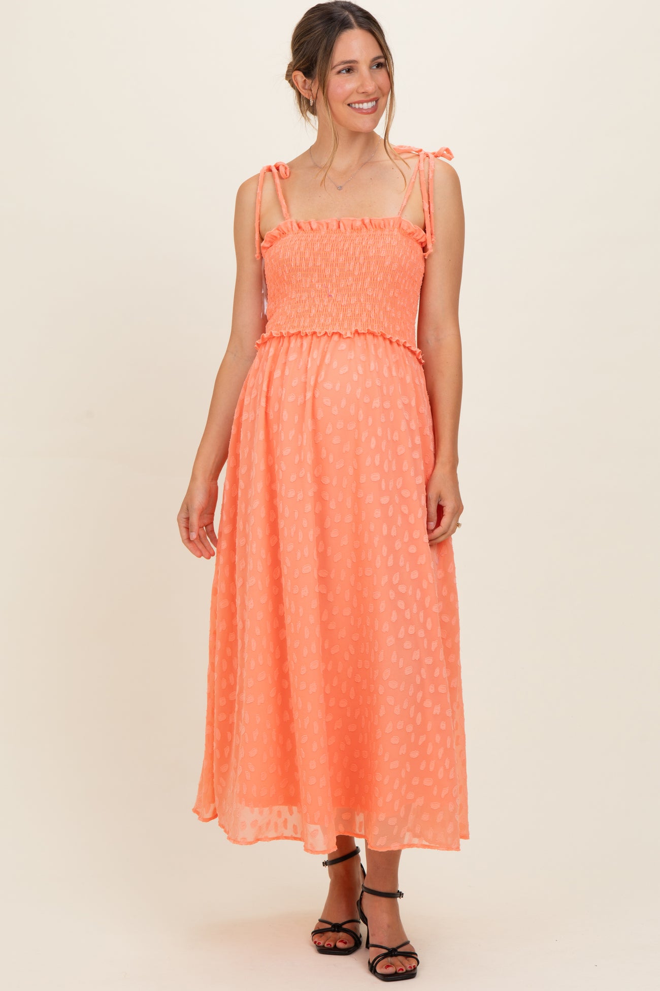 woman wearing a peach-colored maternity dress with tie straps and smocked bodice, showcasing her baby bump