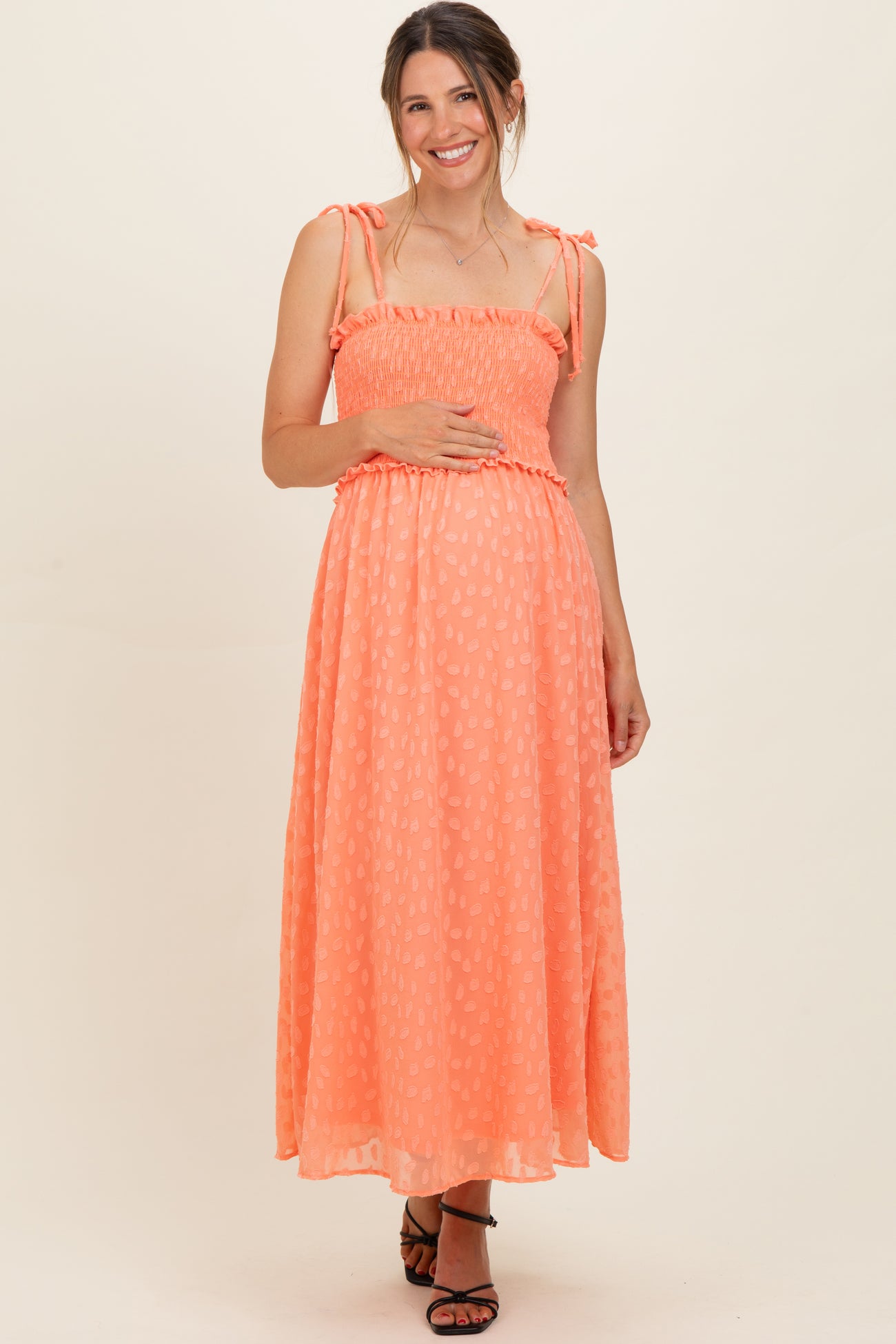 woman wearing a coral maternity maxi dress with adjustable straps, smocked bodice, and holding her baby bump