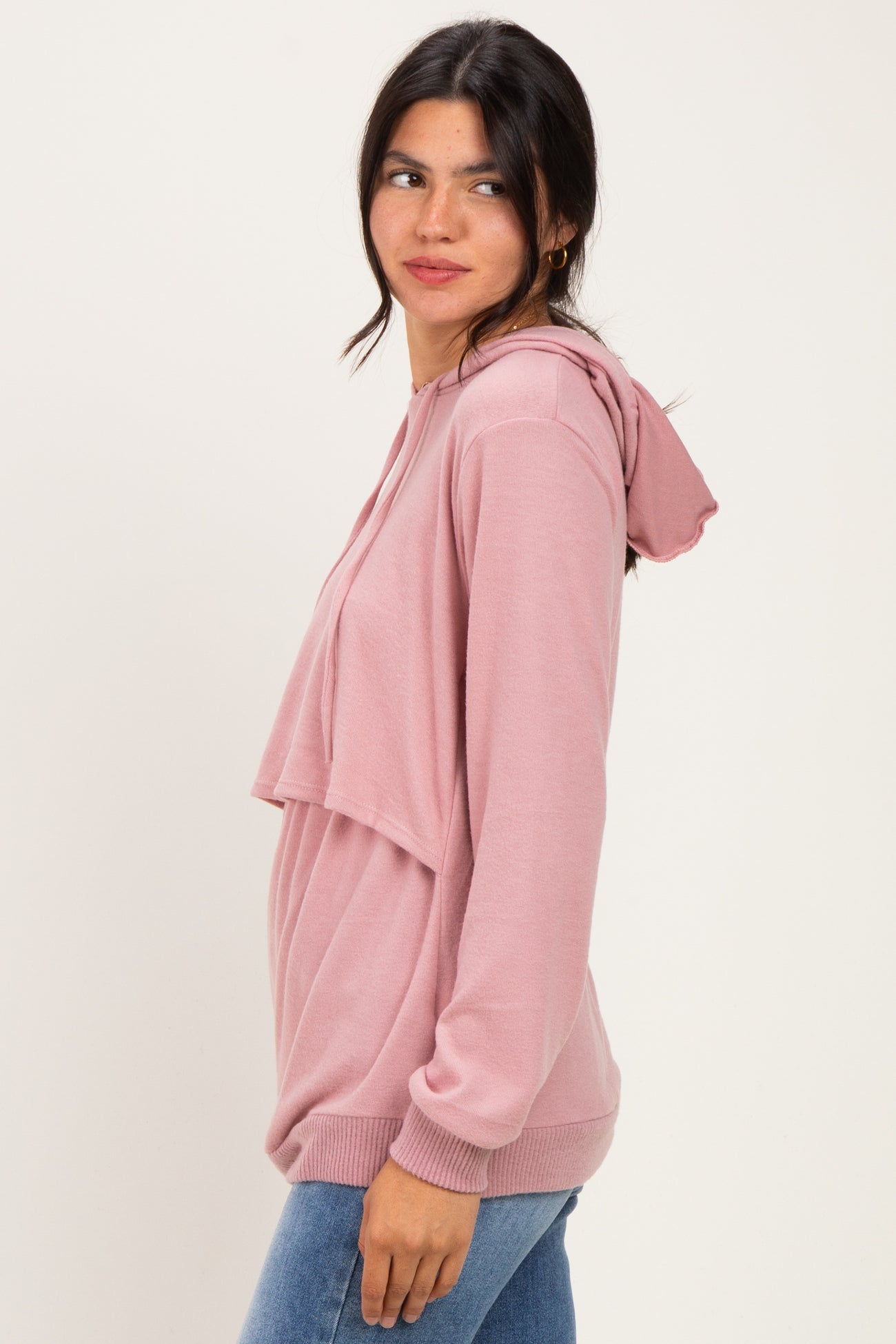 woman wearing a light pink oversized hoodie with a hood and a layered front, standing sideways against a neutral background