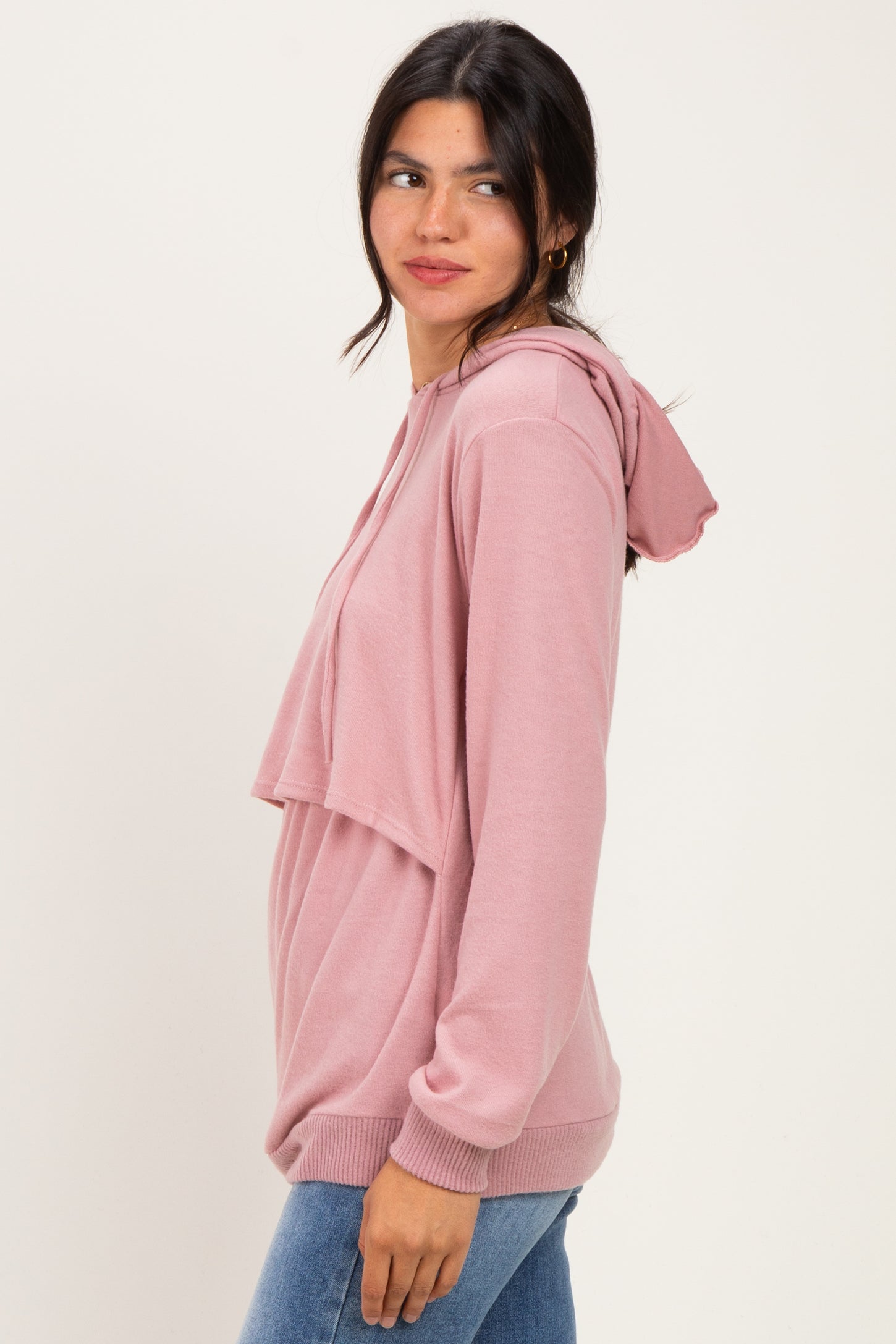 woman wearing a light pink oversized hoodie with a hood and a layered front, standing sideways against a neutral background
