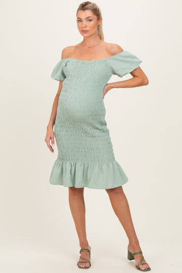 woman wearing a sage green off-the-shoulder maternity dress with smocked details and a ruffled hem, showing her baby bump
