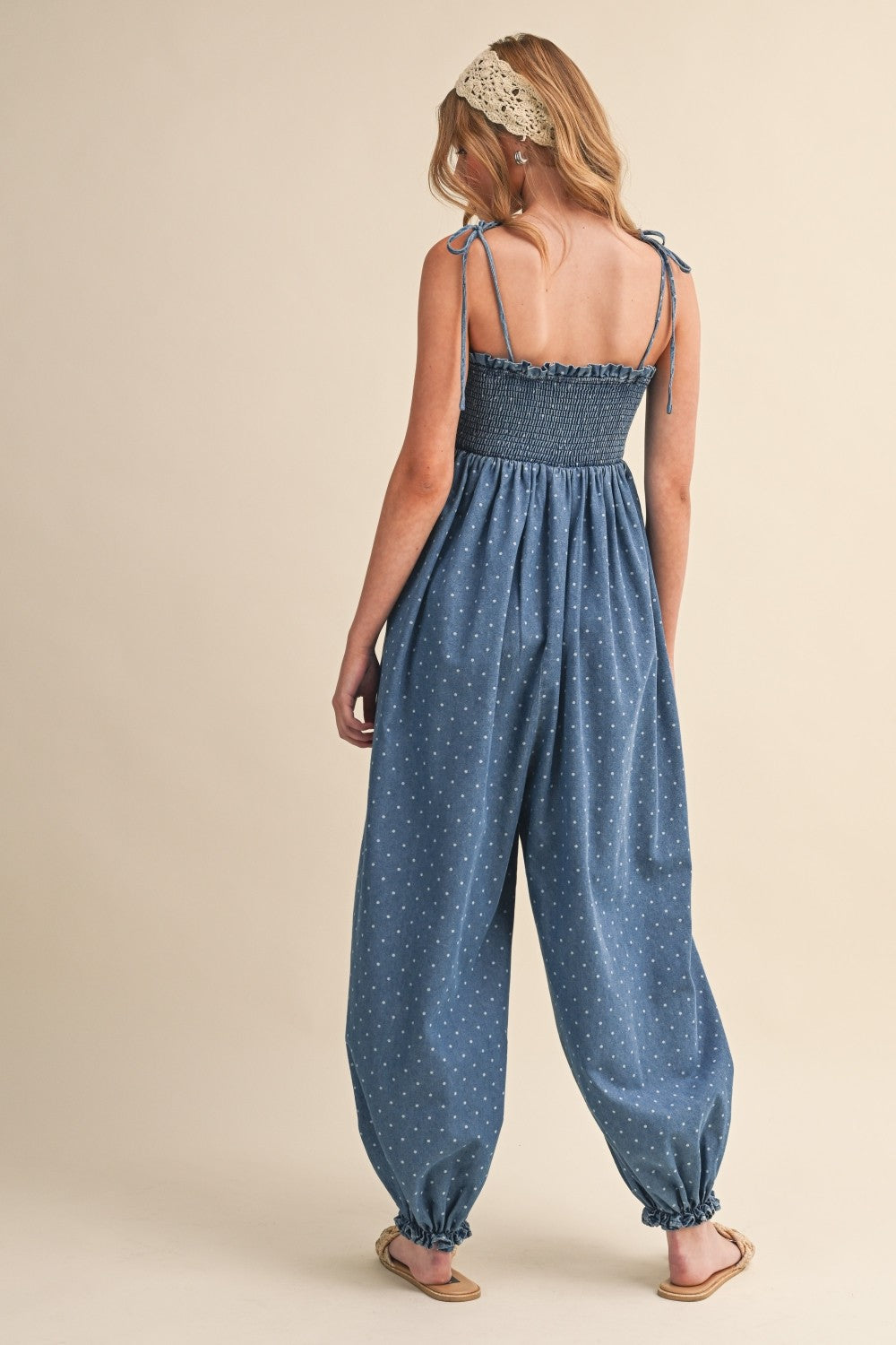 woman wearing a denim blue smocked back jumpsuit with polka dots and adjustable tie straps