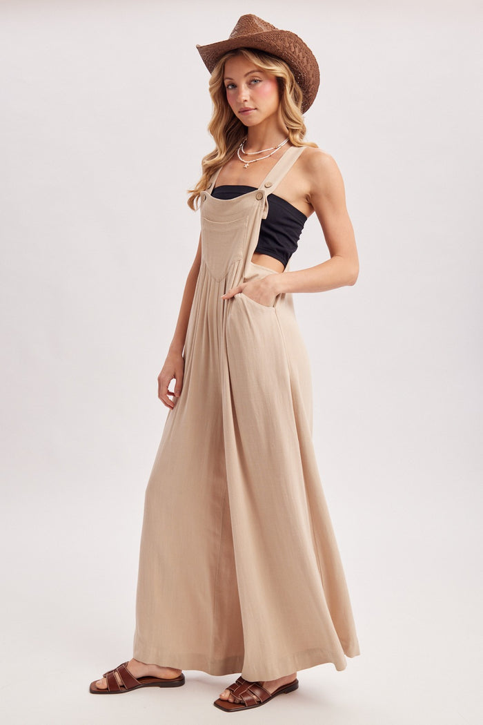 woman wearing a beige long flowy overall dress with pockets, styled with a black tank top and a brown cowboy hat