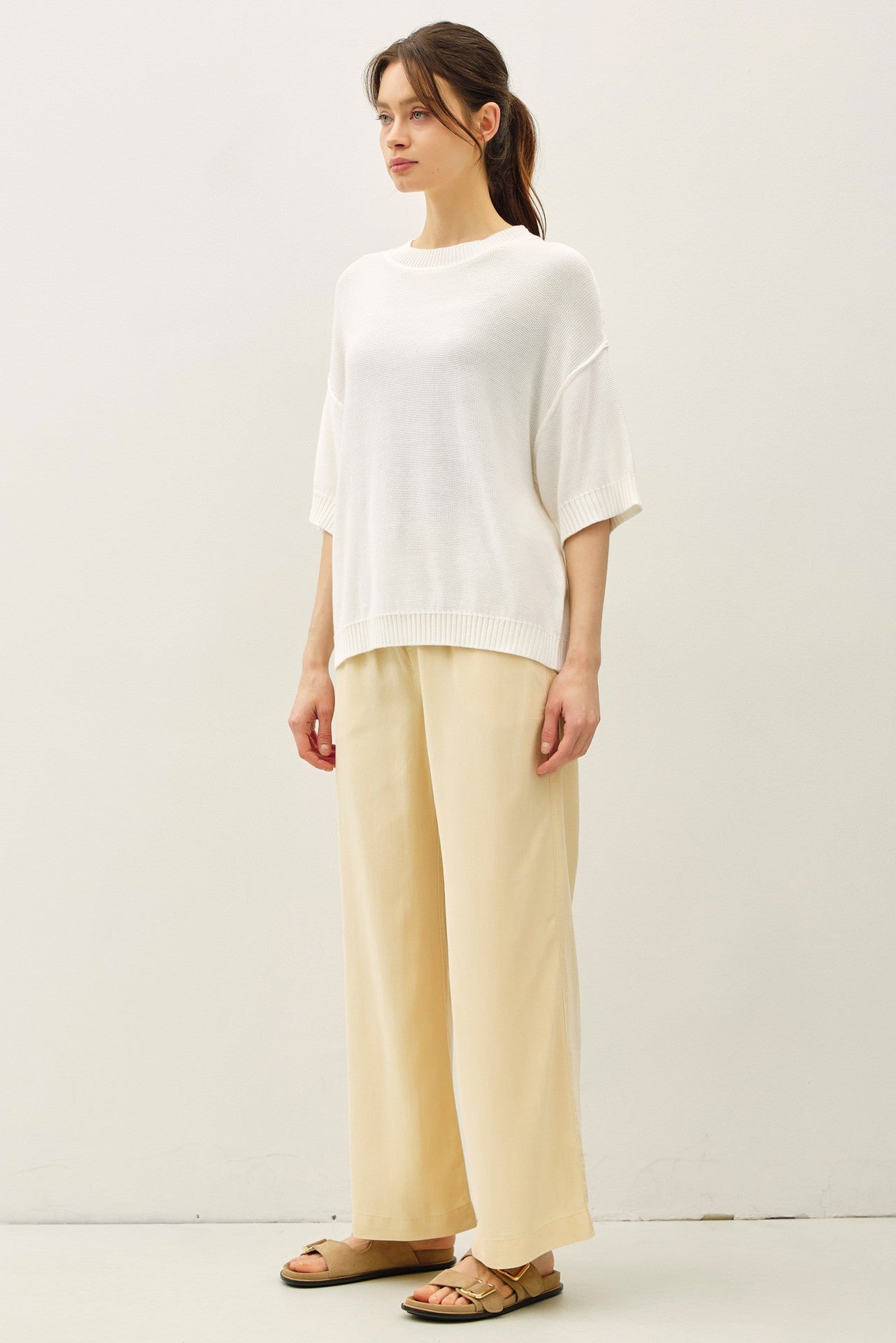 woman wearing a white oversized knit sweater and beige wide-leg trousers, standing against a plain background