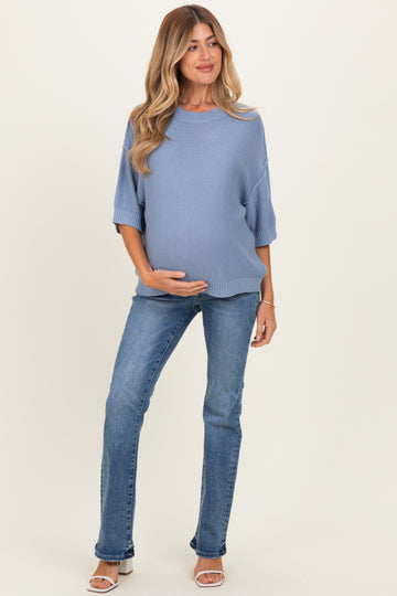 woman wearing a light blue oversized maternity sweater and blue jeans, holding her baby bump