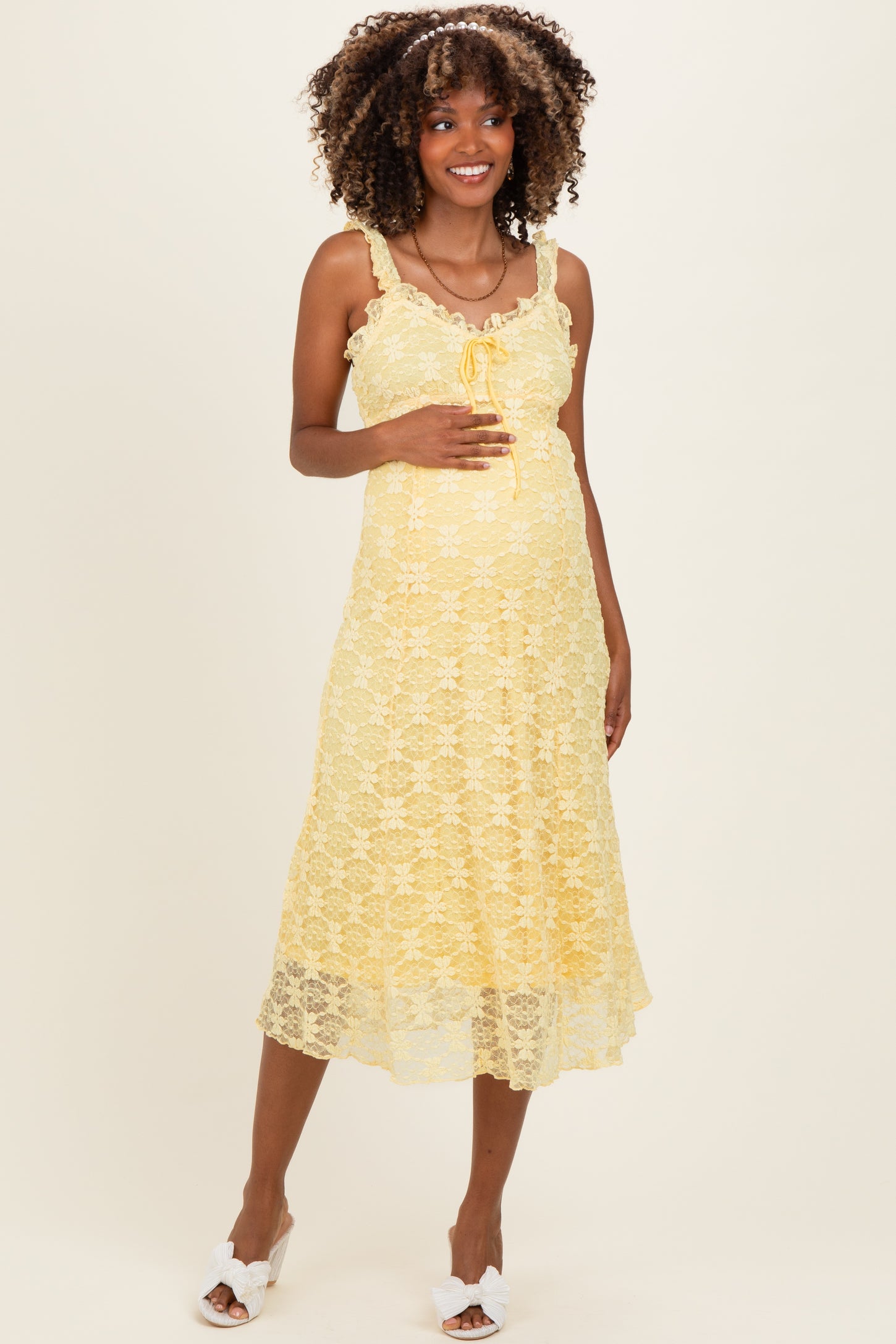woman wearing a yellow lace maternity dress with spaghetti straps and a flowy hem, gently cradling her baby bump