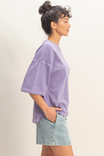 woman wearing a oversized purple t-shirt and light denim shorts, posed in profile with hands in pockets