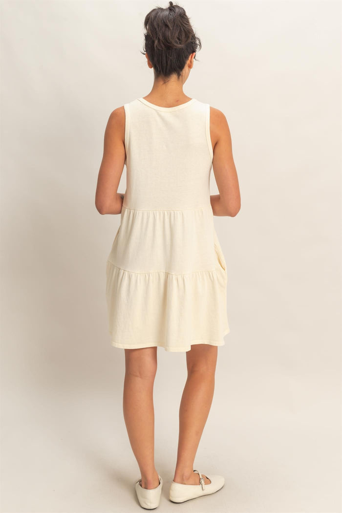 woman wearing a sleeveless cream tiered dress, showing the back view with a relaxed fit and a flared hem