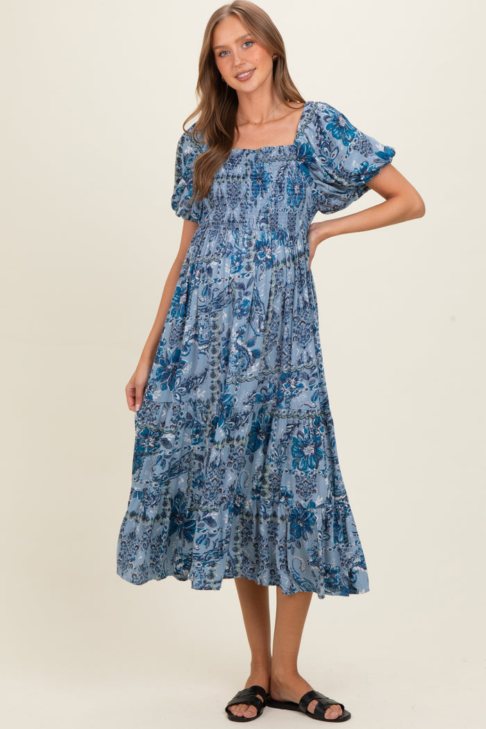 woman wearing a blue floral print maternity dress with puff sleeves and a flowing skirt, smiling with one hand on her hip