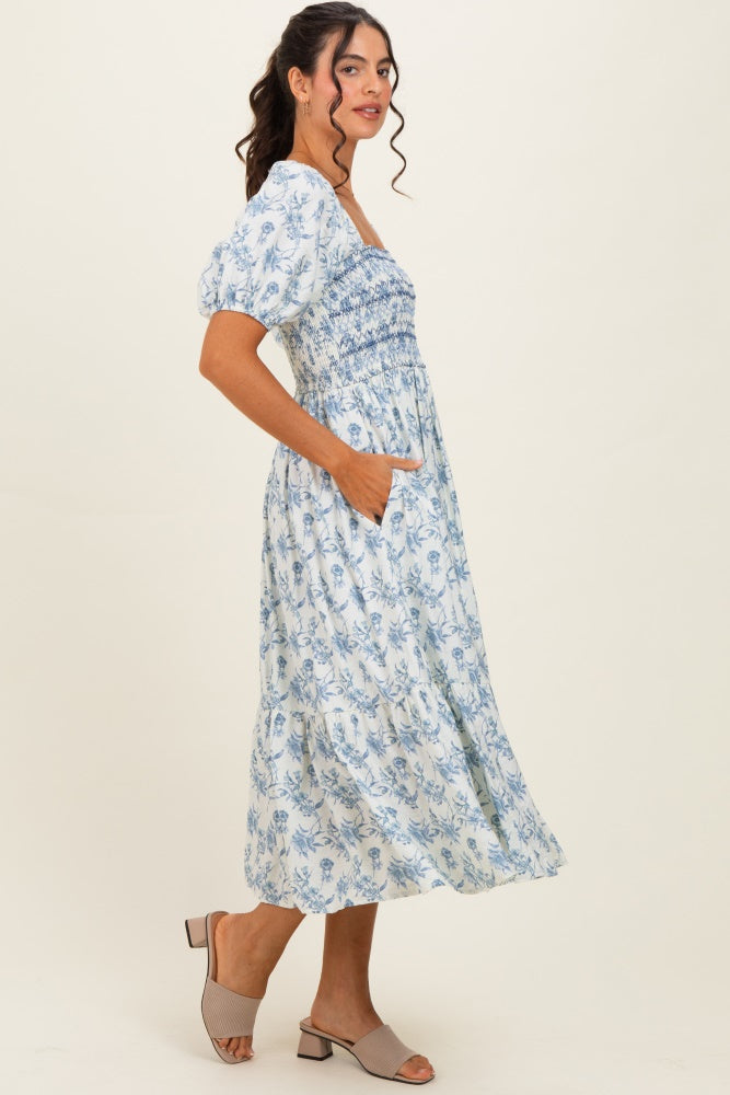 woman wearing a blue and white floral smock dress with puff sleeves and pockets, standing sideways