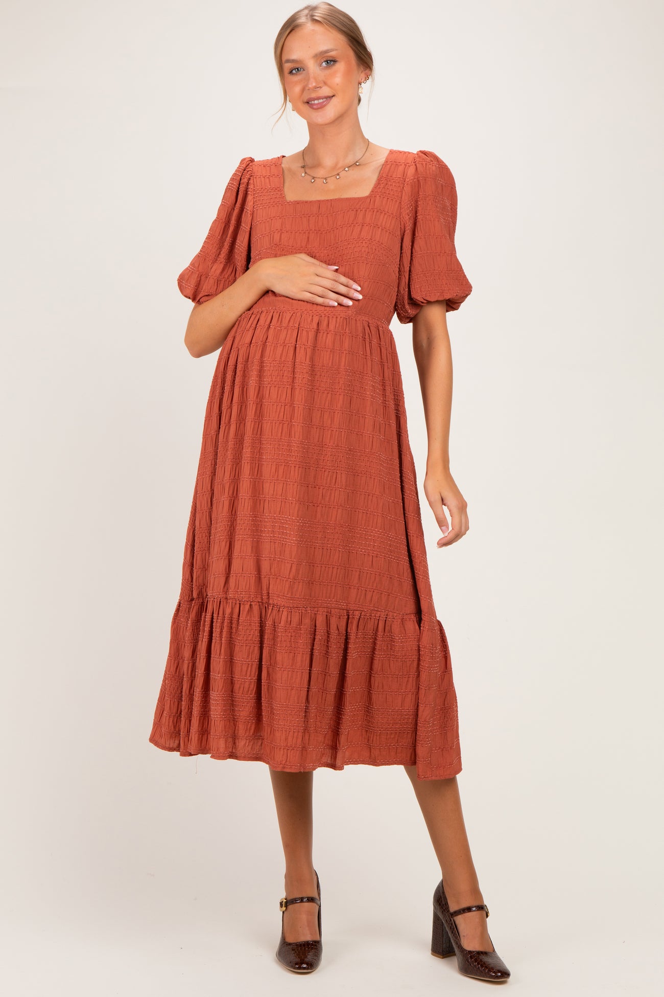 woman wearing a rust-colored short sleeve maternity dress with a square neckline and tiered skirt, holding her baby bump
