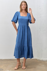 woman wearing a blue midi dress with square neckline and puff sleeves, standing against a white wall