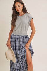 woman wearing a gray cropped t-shirt and a blue plaid maxi skirt, holding a beige hat and posing with one leg visible