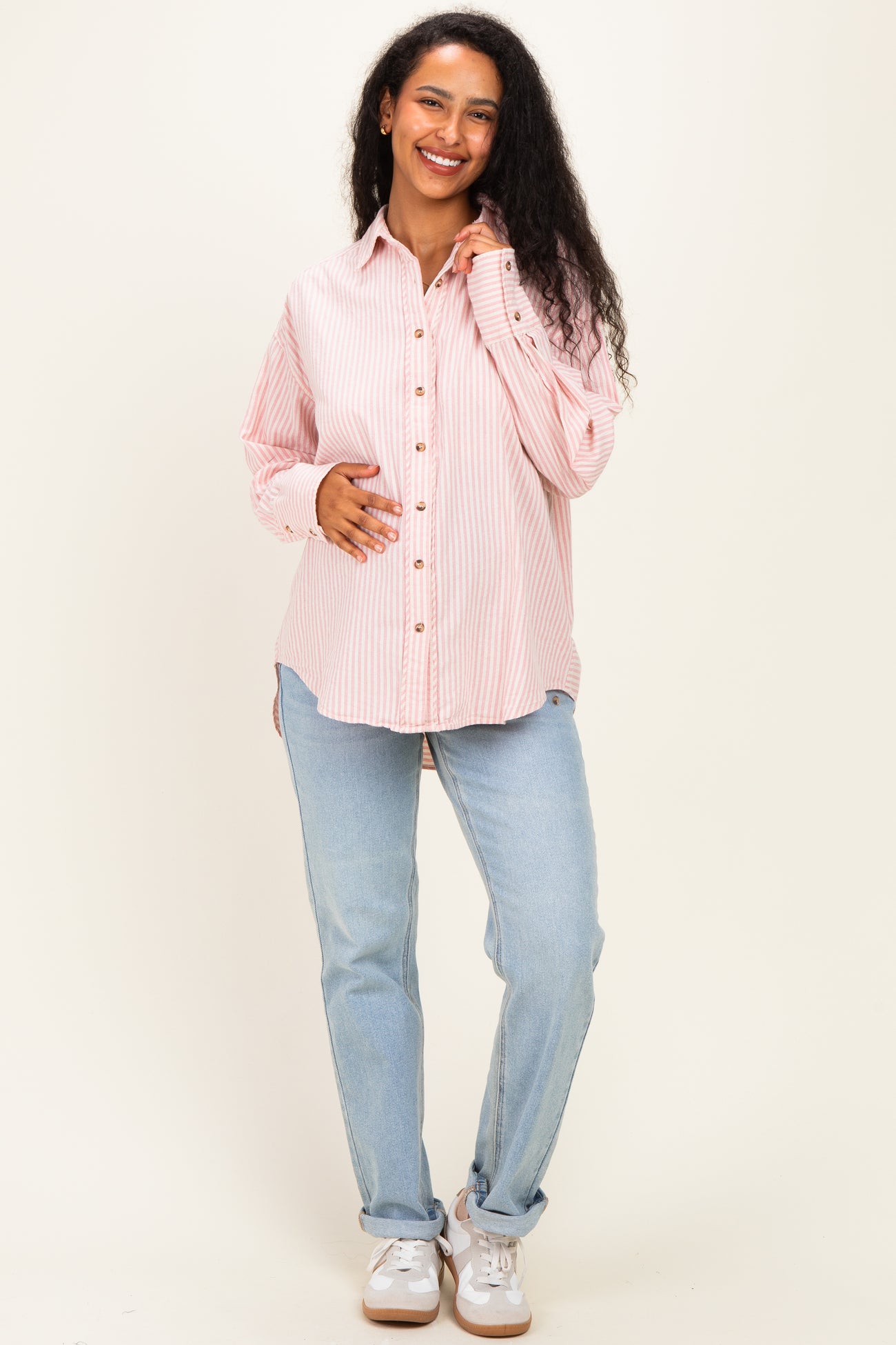 woman wearing a pink and white striped maternity button-up shirt and light blue jeans, cradling her baby bump