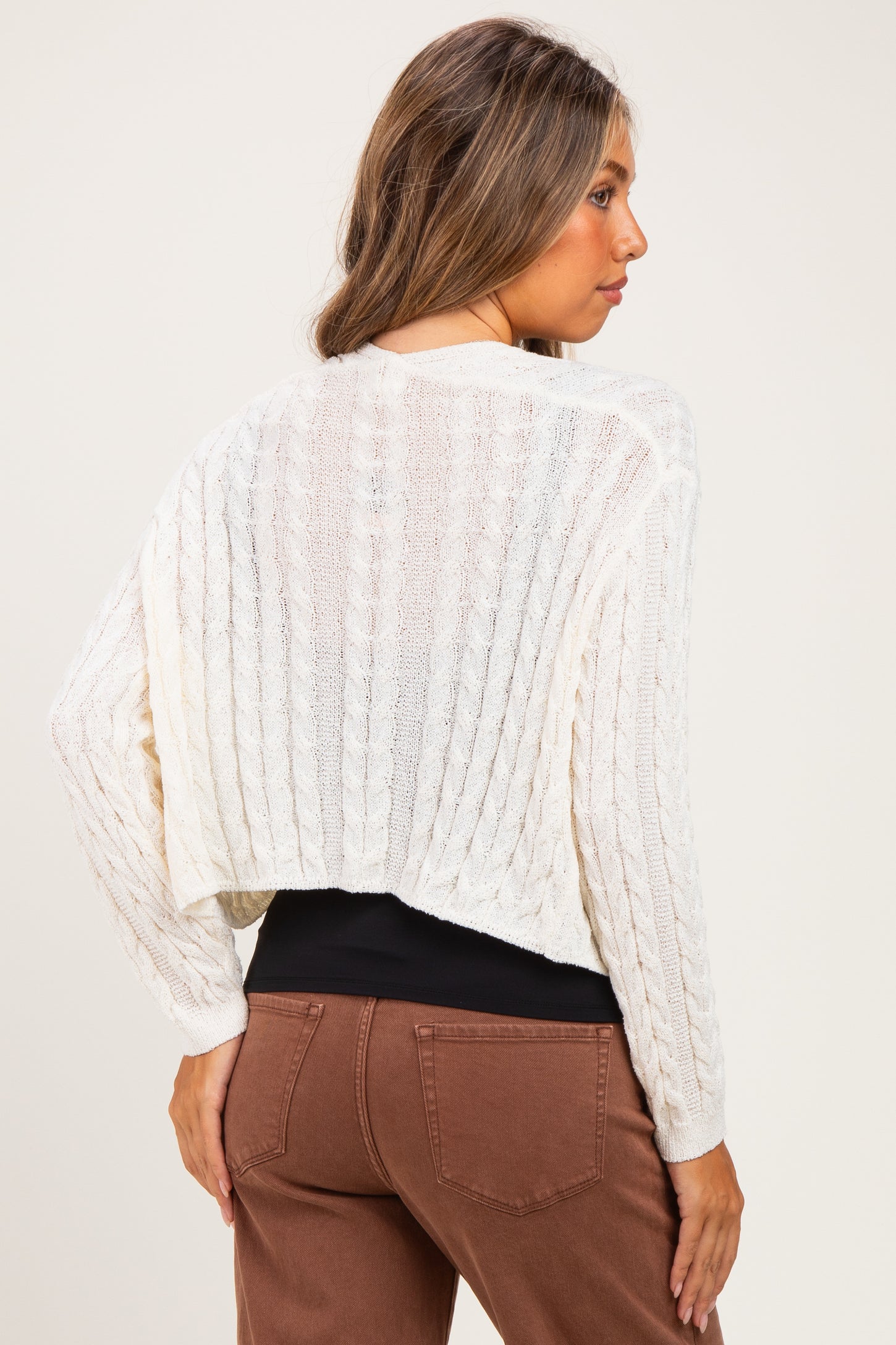 woman wearing a cream cable knit maternity sweater with a cropped back, styled with brown pants
