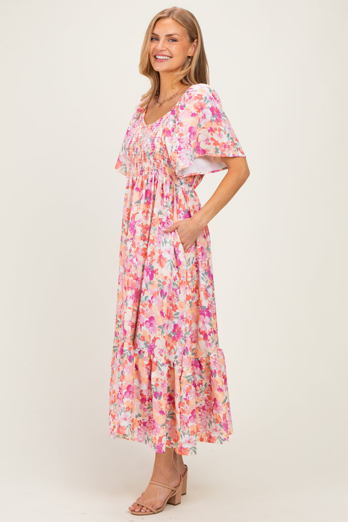 woman wearing a floral pink maxi dress with short sleeves and pockets, standing and smiling on a neutral background