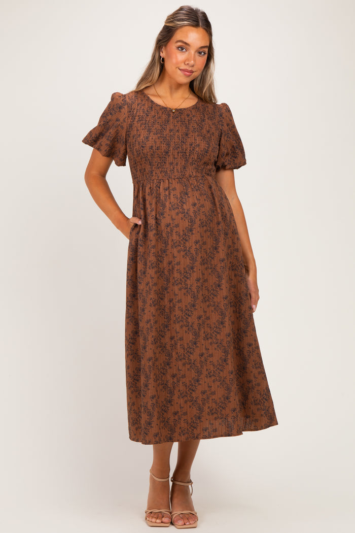 woman wearing a brown floral print maternity dress with short puff sleeves and a smocked bodice, standing with hands in pockets