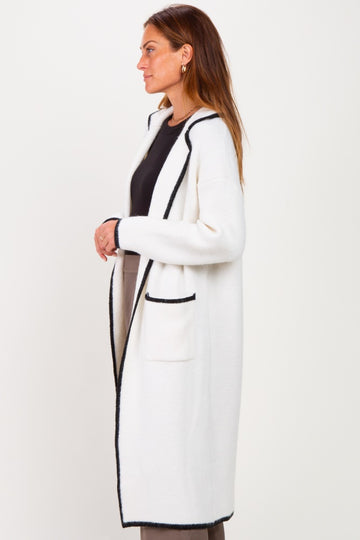 woman wearing a long white cardigan with black trim and pockets, standing sideways against a white background