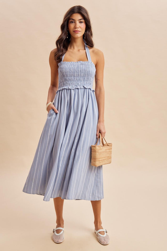 woman wearing a light blue striped halter dress with a smocked bodice and a full skirt, holding a small woven bag