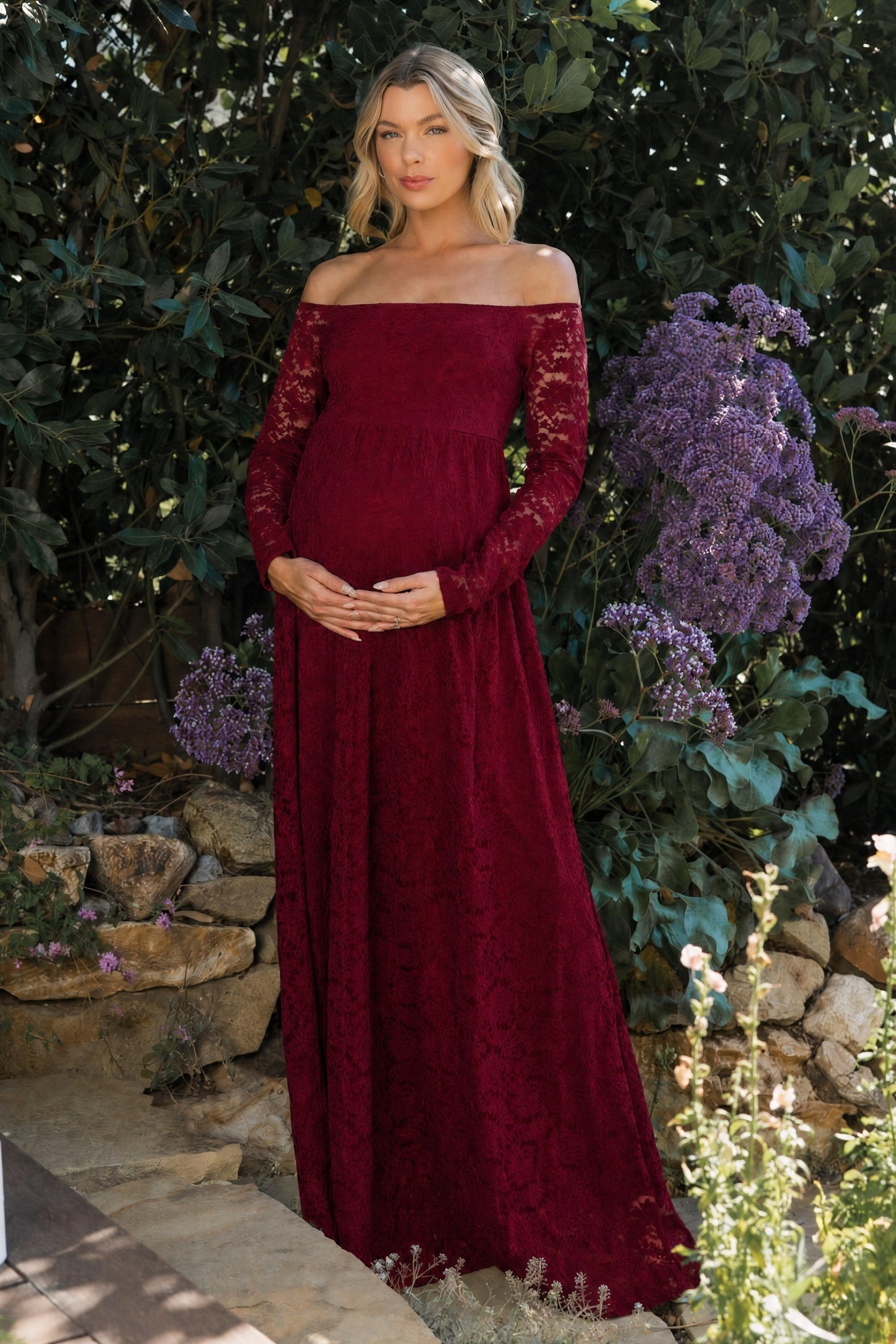 woman wearing a burgundy off-the-shoulder lace maternity dress and cradling her baby bump in a garden setting
