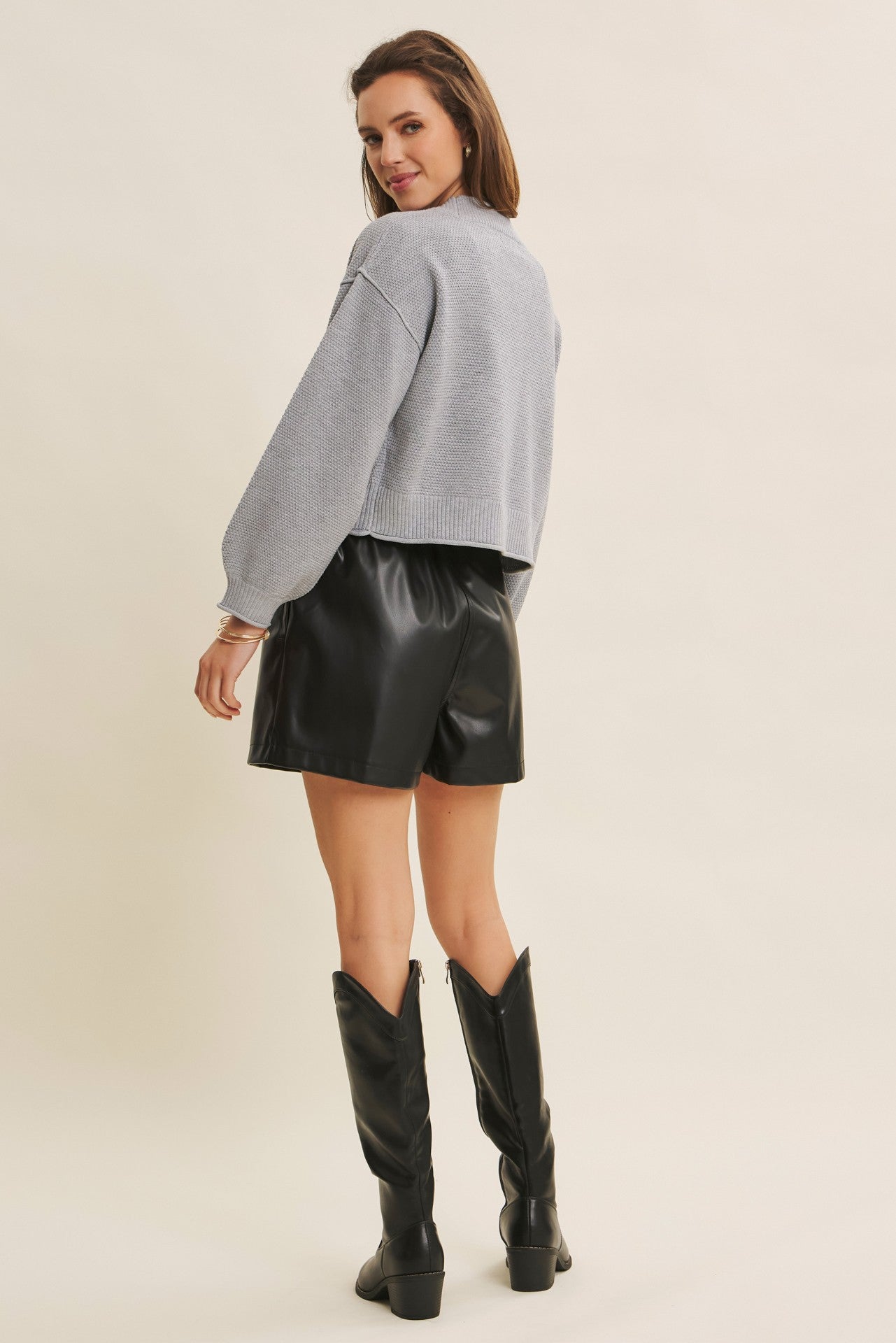 woman wearing a light gray cropped sweater and black faux leather shorts, standing with her back to the camera