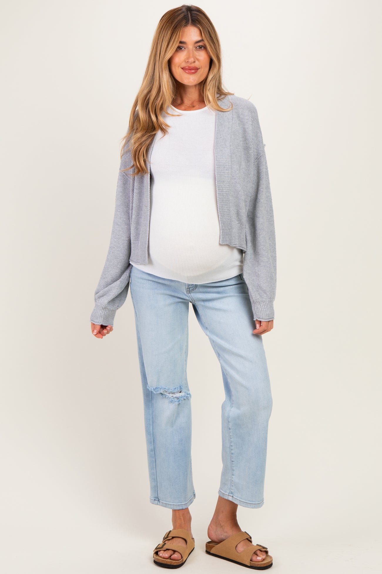 woman wearing a light gray maternity cardigan over a white tank top and blue distressed maternity jeans, showing a baby bump