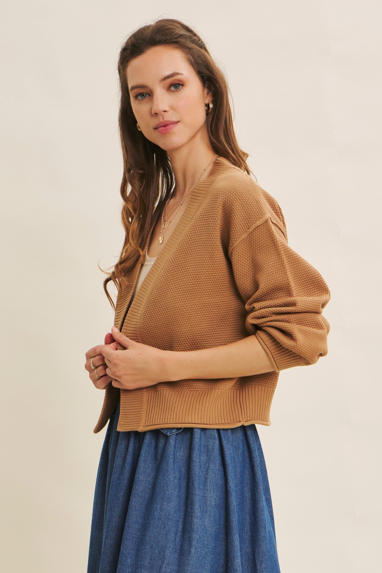 woman wearing a tan cropped cardigan over a cream top and a denim skirt, standing in a side profile pose