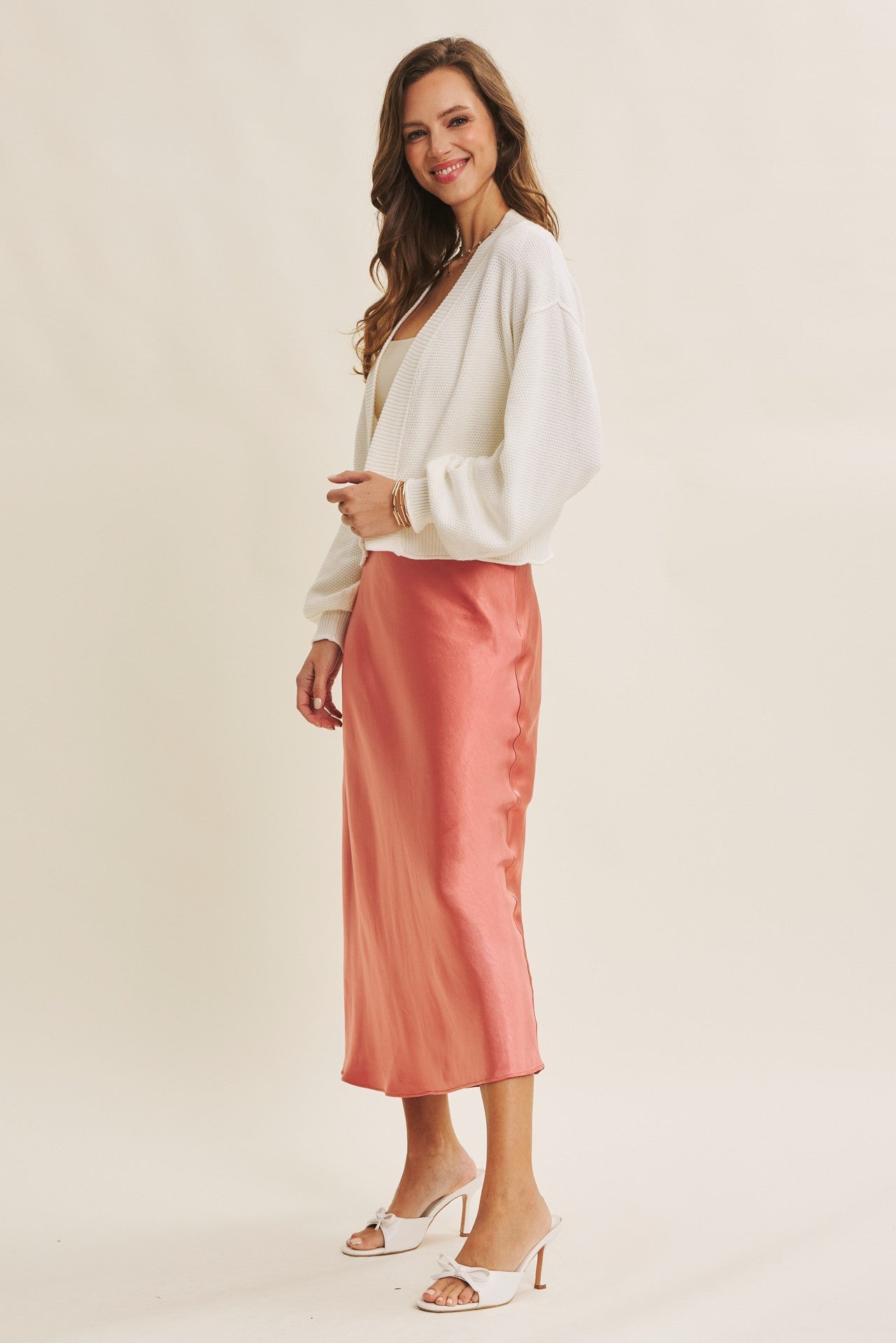 woman wearing a white oversized cardigan and a pink satin midi skirt with heels, smiling to the side
