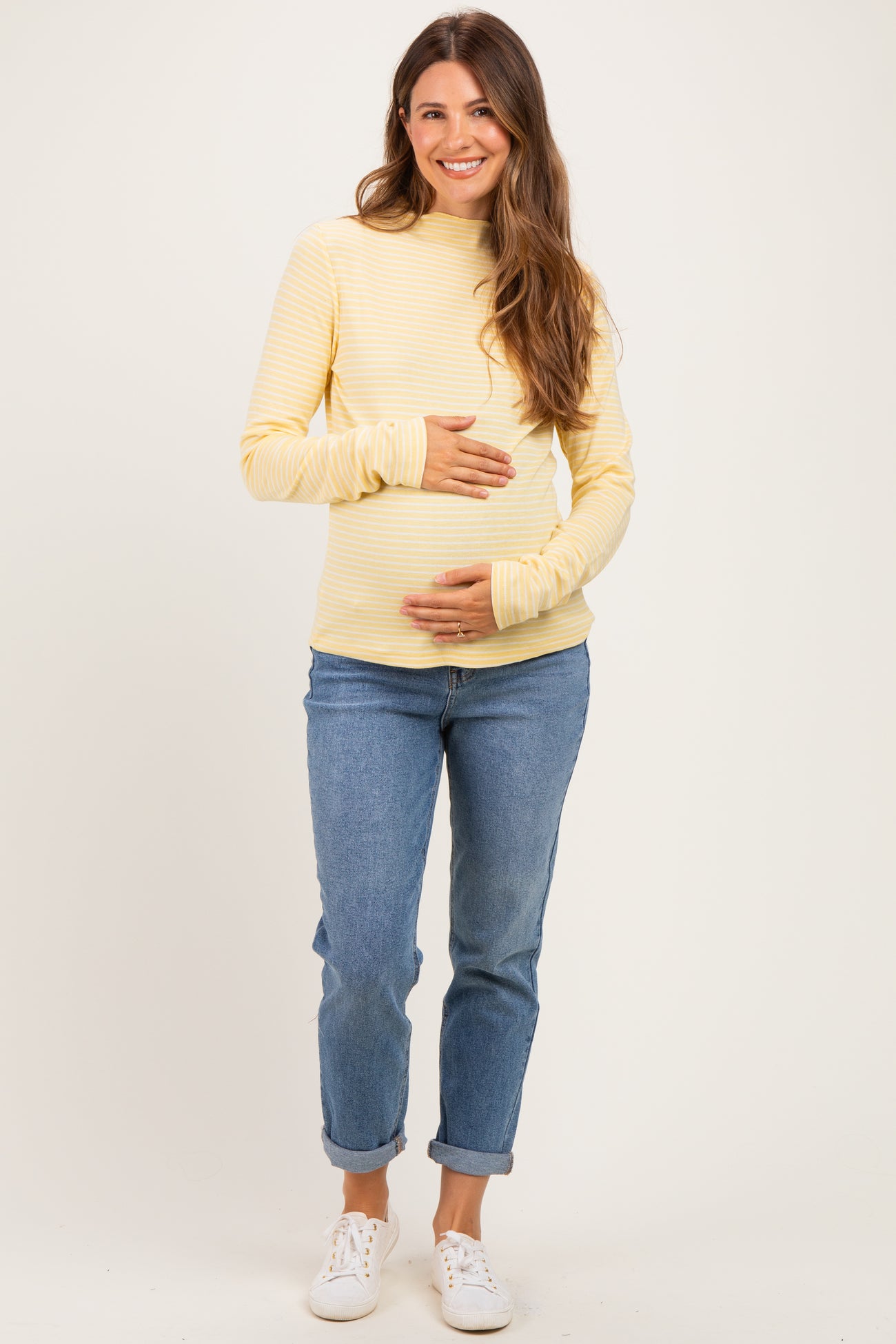 woman wearing a yellow striped long sleeve maternity top and blue jeans, holding her baby bump