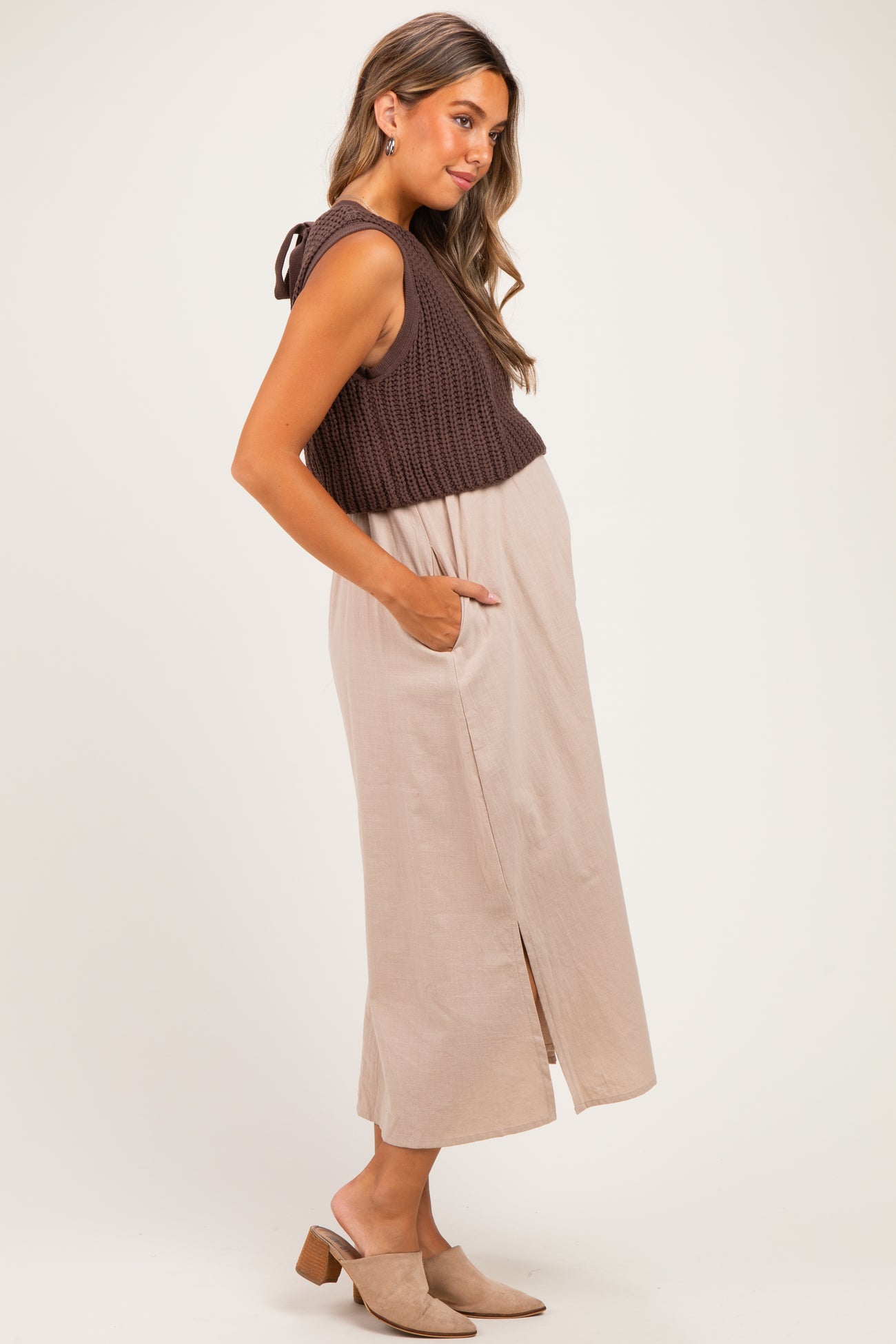 woman wearing a brown knitted maternity crop top and a beige maxi dress with side pockets and a front slit, showing her baby bump