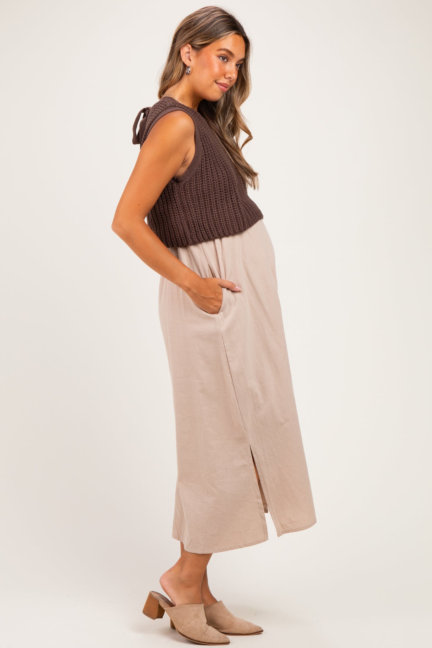 woman wearing a brown knitted maternity crop top and a beige maxi dress with side pockets and a front slit, showing her baby bump