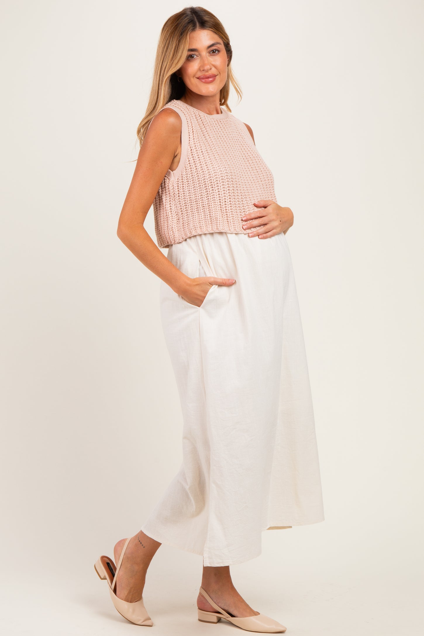 woman wearing a sleeveless peach maternity top and a white maxi skirt, holding her baby bump.