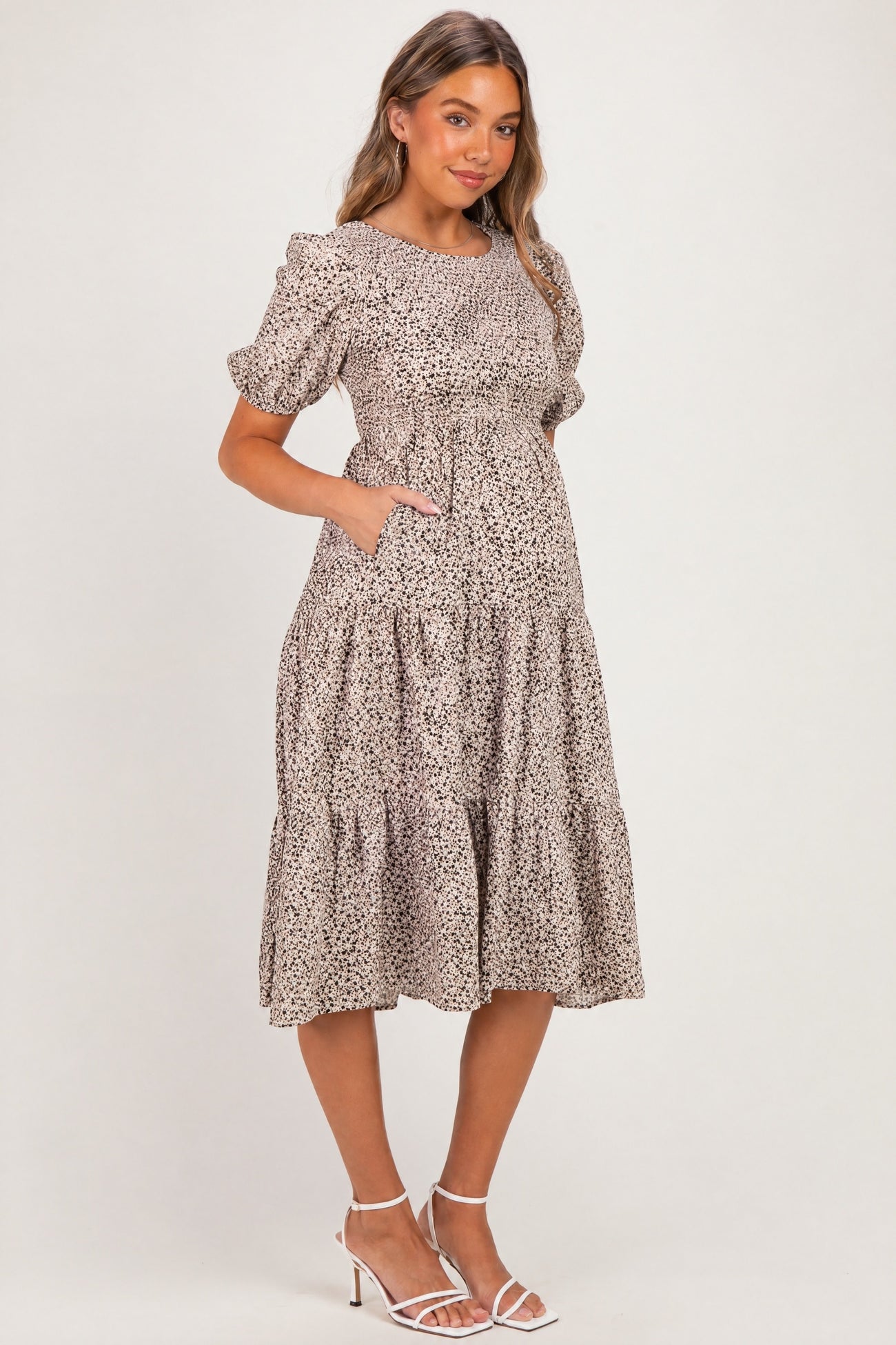 woman wearing a beige floral print maternity dress with short puff sleeves and pockets, standing with a baby bump visible